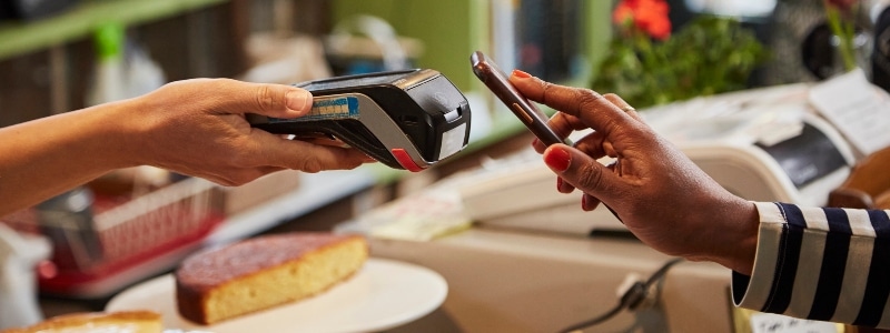 A hand holding a phone reaches out to pay with contactless payment, while another hand reaches out with a payment processer.