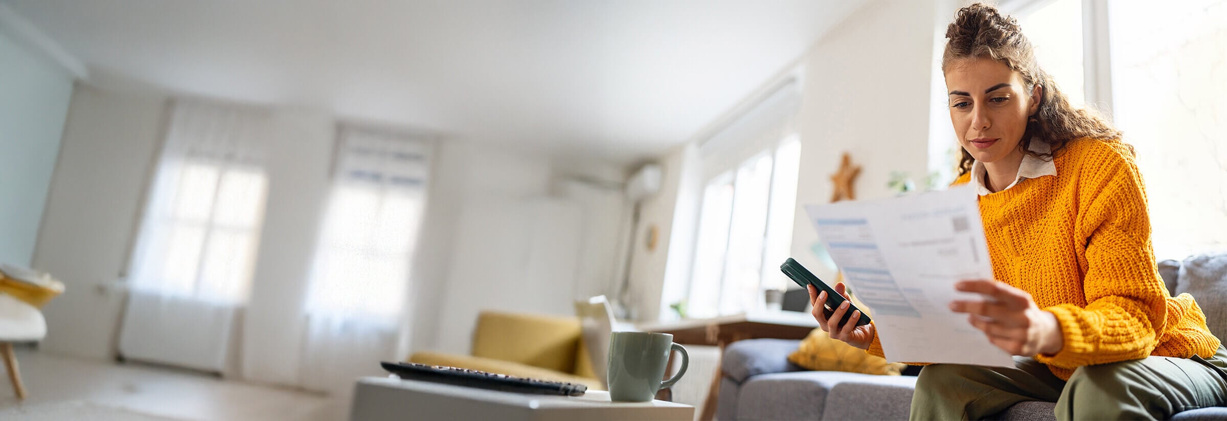 A woman looking at a sheet of paper while checking if her credit score has updated on her smartphone.