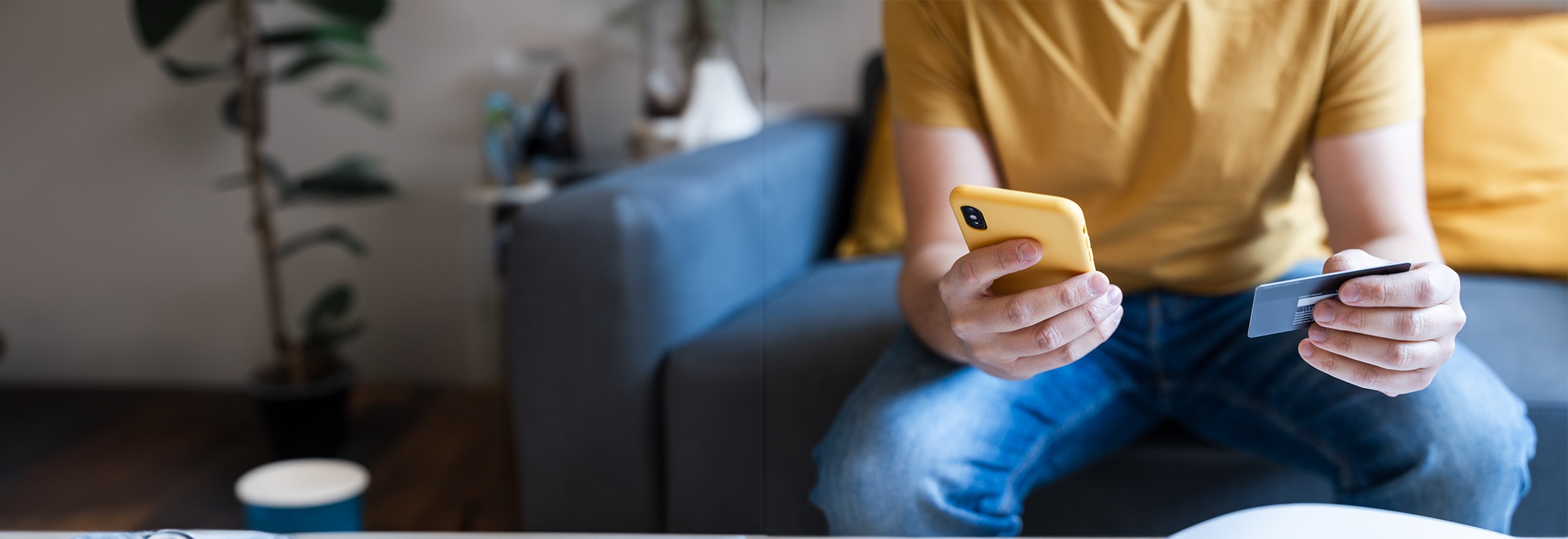 Person holding a yellow smartphone in one hand and a credit card in the other, seated on a couch.