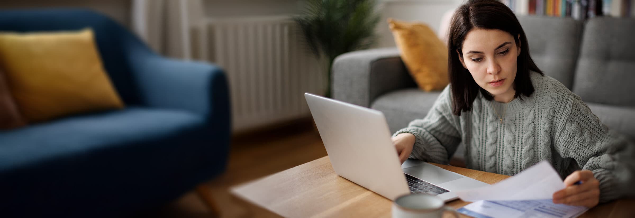 A woman in a gray sweater works on a laptop and looks at papers, potentially doing her taxes.