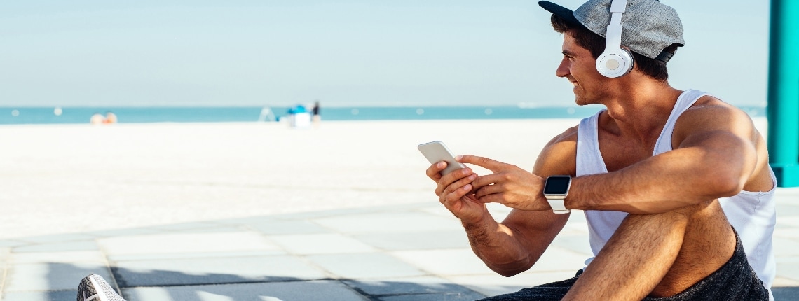 A person sitting down, wearing headphones, and holding a mobile device by a beach