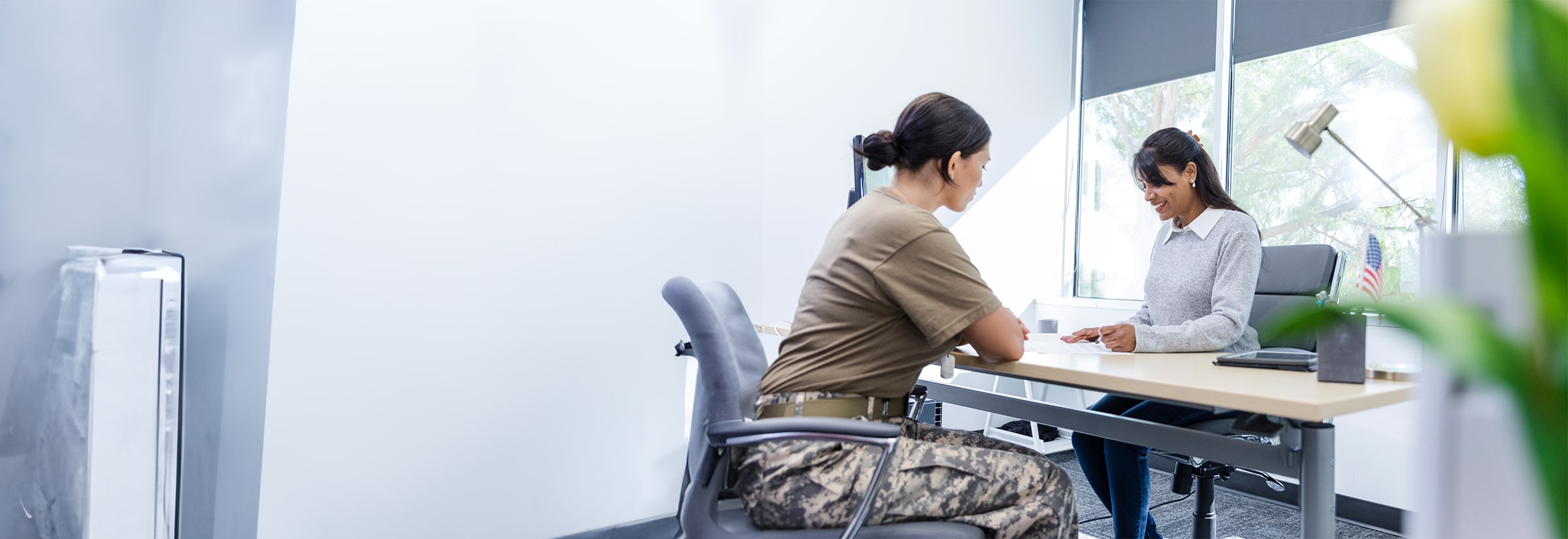 Two people sitting at a desk. One woman is dressed in military attire.