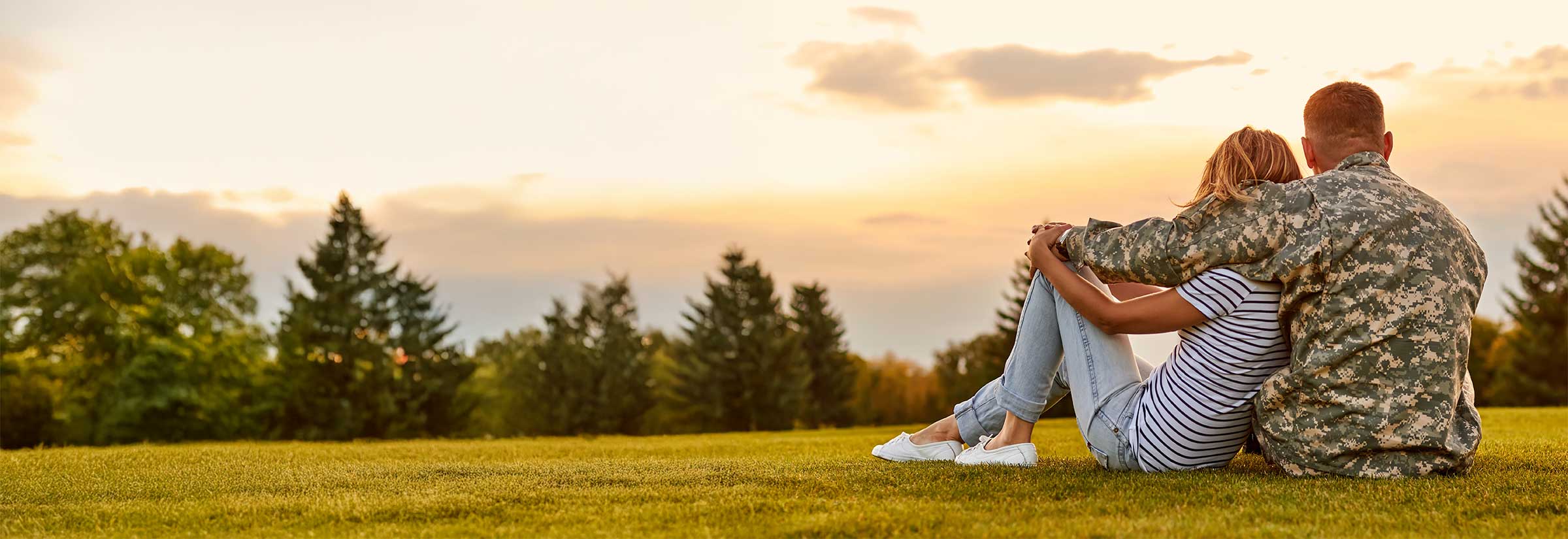 A woman and a man in military uniform sit in a field, romantically embracing as they watch the sunset.