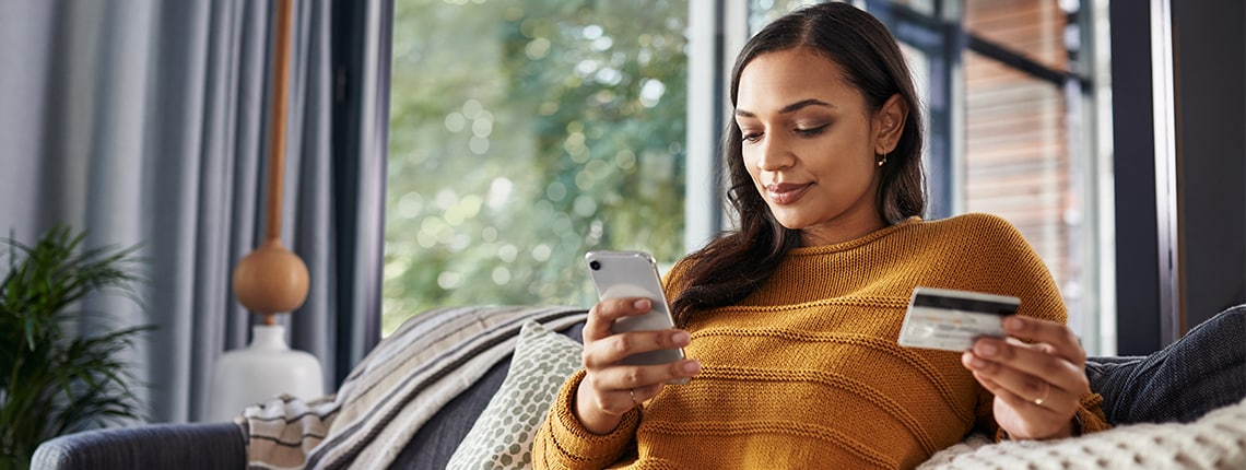 A woman sitting on a couch holding a mobile device and credit card.