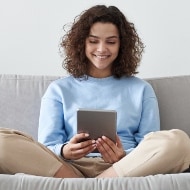 A woman sits on a grey couch with her legs crossed, looking down at her device.